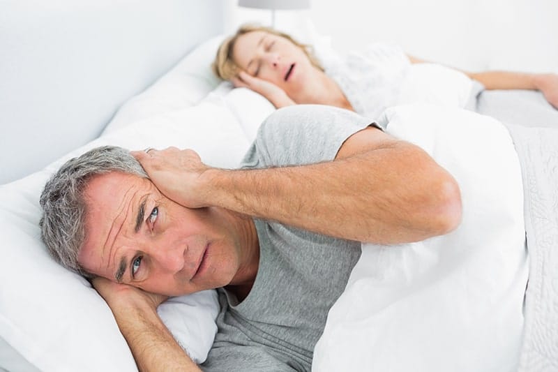 man covering his ears in bed while wife snores next to him