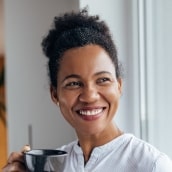 services-implants-min Woman smiling and holding a coffee cup, showcasing dental implants with bright, even teeth.