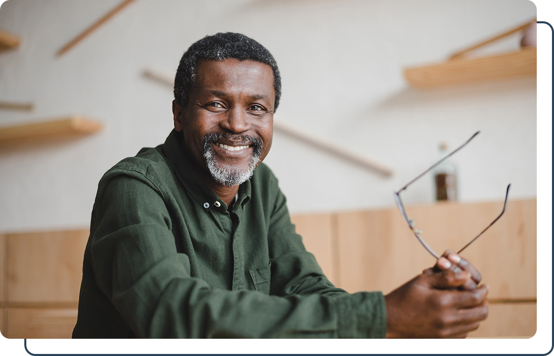 Kuhn Man smiling holding his glasses Smiling man in a green shirt holding glasses, seated in a modern room with wooden shelves and light decor.