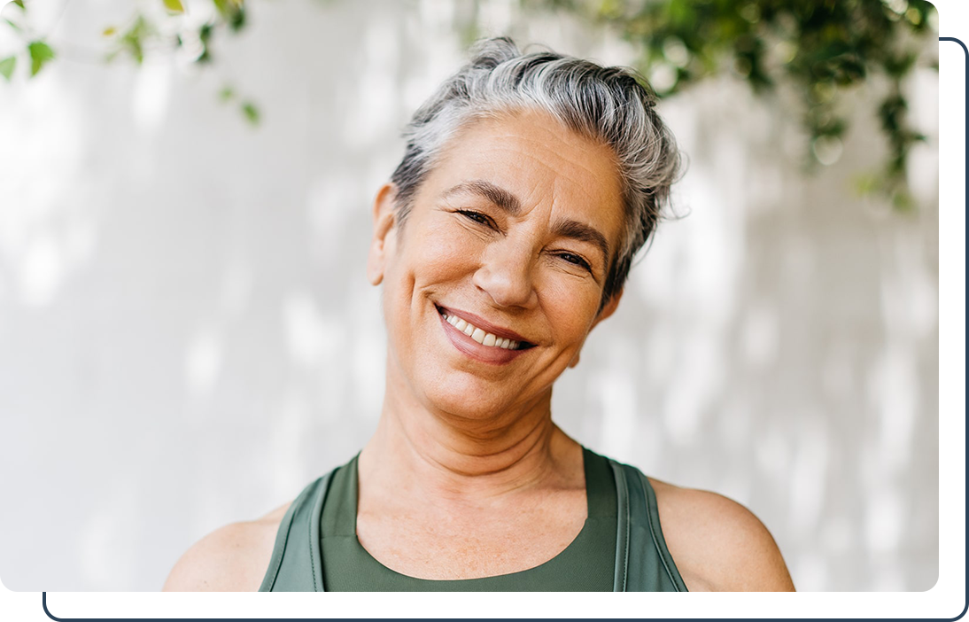Kuhn woman smiling in athletic wear A woman with short gray hair smiling warmly, wearing a green top, standing outdoors with soft greenery in the background.